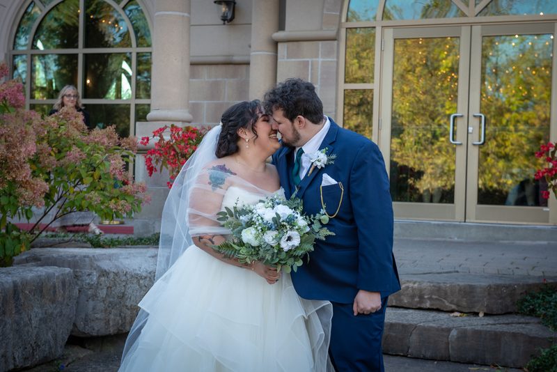 A bride in a white gown and veil kisses her groom in front of stone steps, autumn foliage, and festive string lights.