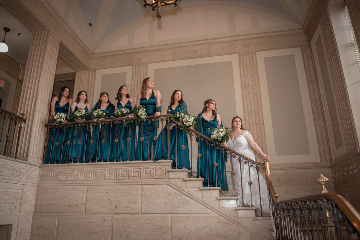 A bride in a white gown descends a marble staircase flanked by bridesmaids in teal dresses holding bouquets.