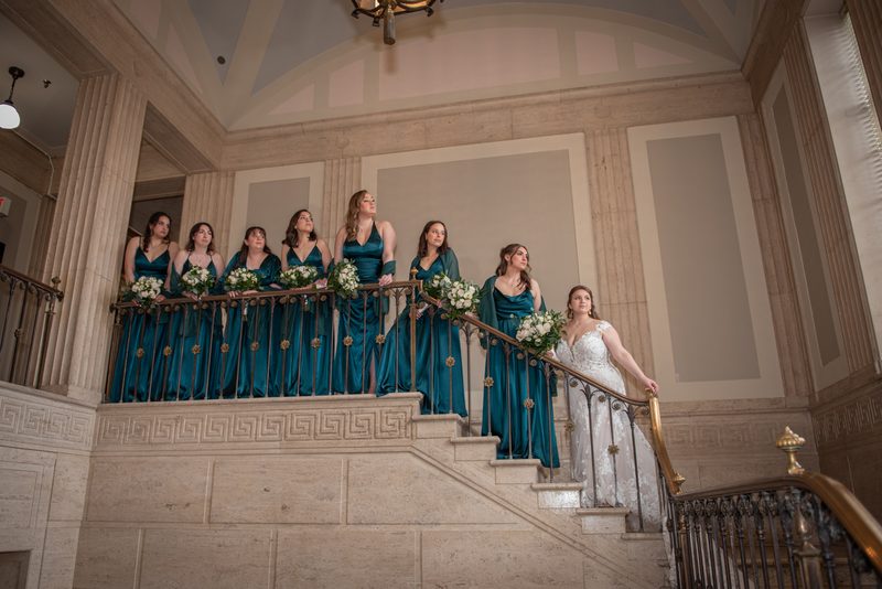 A bride in a white gown descends a marble staircase flanked by bridesmaids in teal dresses holding bouquets.