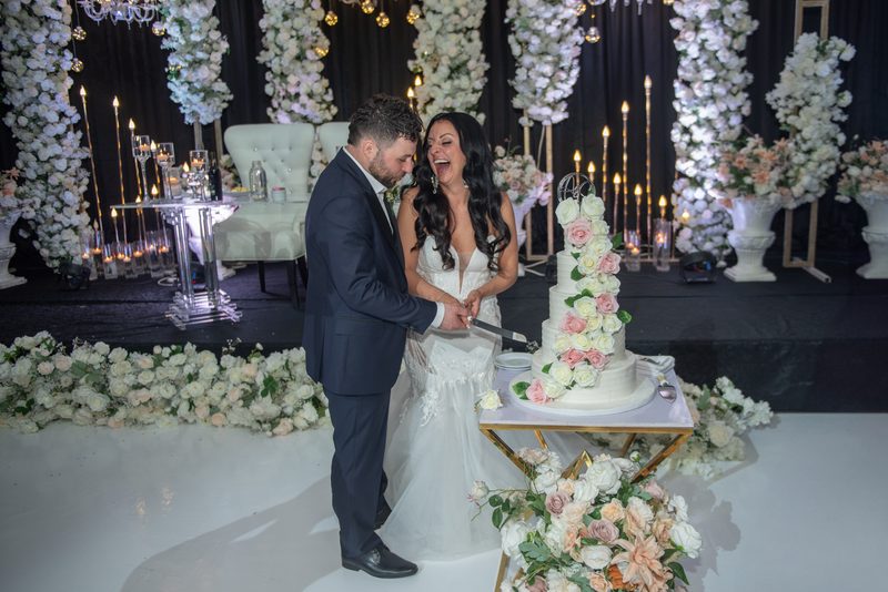Bride and groom laugh as they cut their multi-tiered wedding cake, surrounded by candles and floral arrangements.
