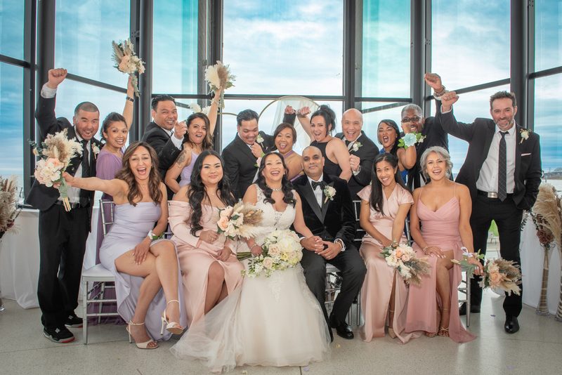 Bride and groom surrounded by joyful guests raising fists indoors near large windows with cloudy sky visible.