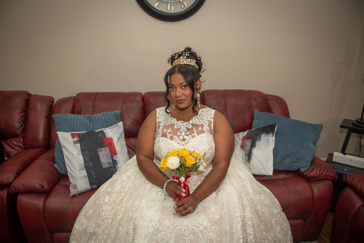 A young woman in a white lace quinceañera gown and tiara holds flowers while sitting on a red leather couch.