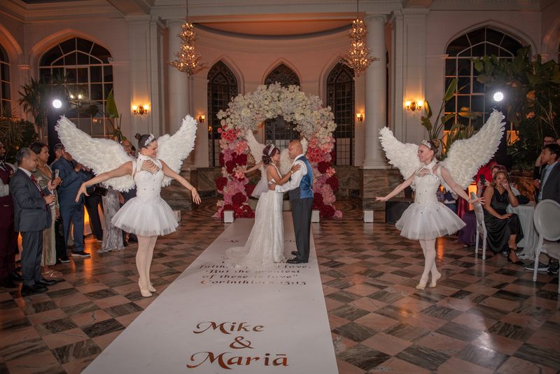 Bride and groom stand at altar as ballerinas in angel costumes dance beside them, guests watch from sides.