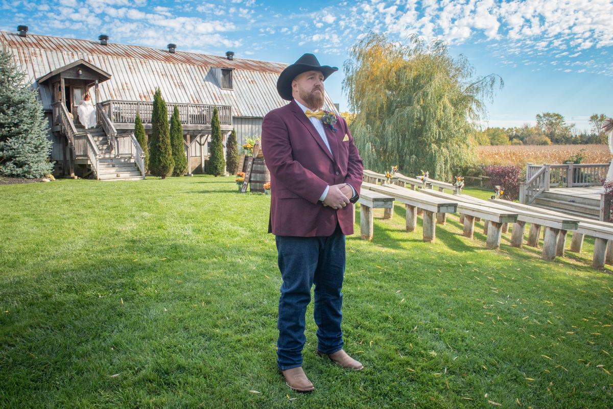 A groom in a maroon blazer and yellow bow tie stands on grass, watching the bride descend wooden stairs from a rustic barn.