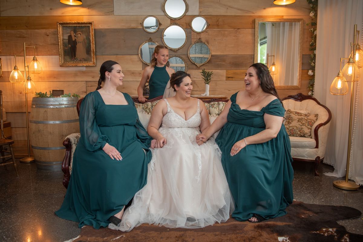 A bride in white sits with two laughing bridesmaids in teal dresses in an intimate, warmly lit room.