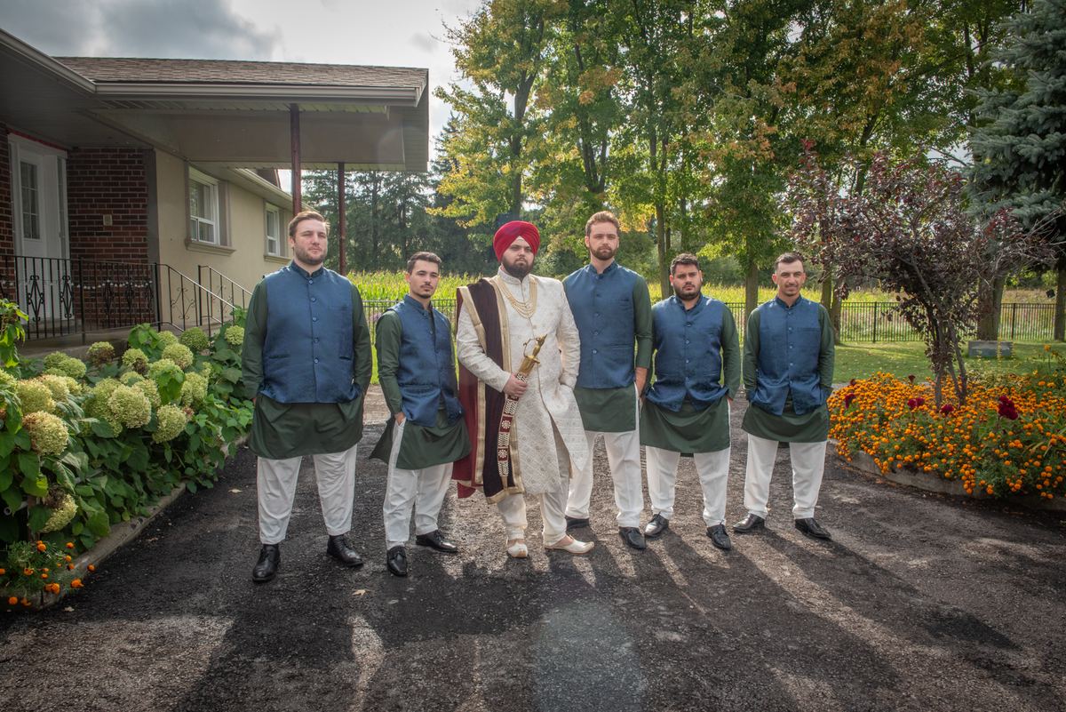 A Sikh groom in red turban and sword stands with four groomsmen beside a house, posing outdoors on a paved path.