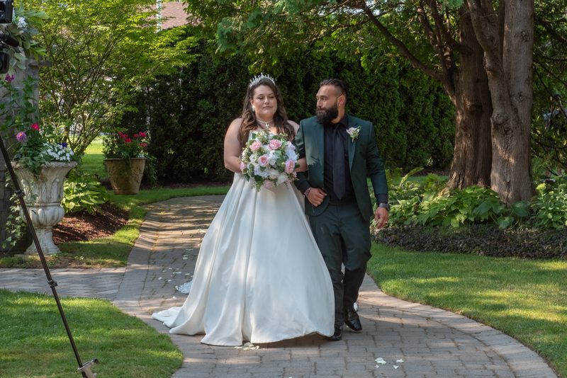 A bride in a white gown and groom in a dark green suit walk hand-in-hand along a stone pathway in a garden.
