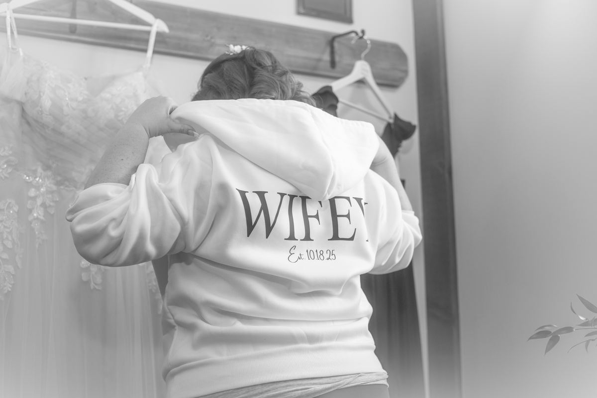 A bride adjusts her white hoodie with 'WIFEY Est. 101825' while standing near a wedding dress, preparing for the ceremony.