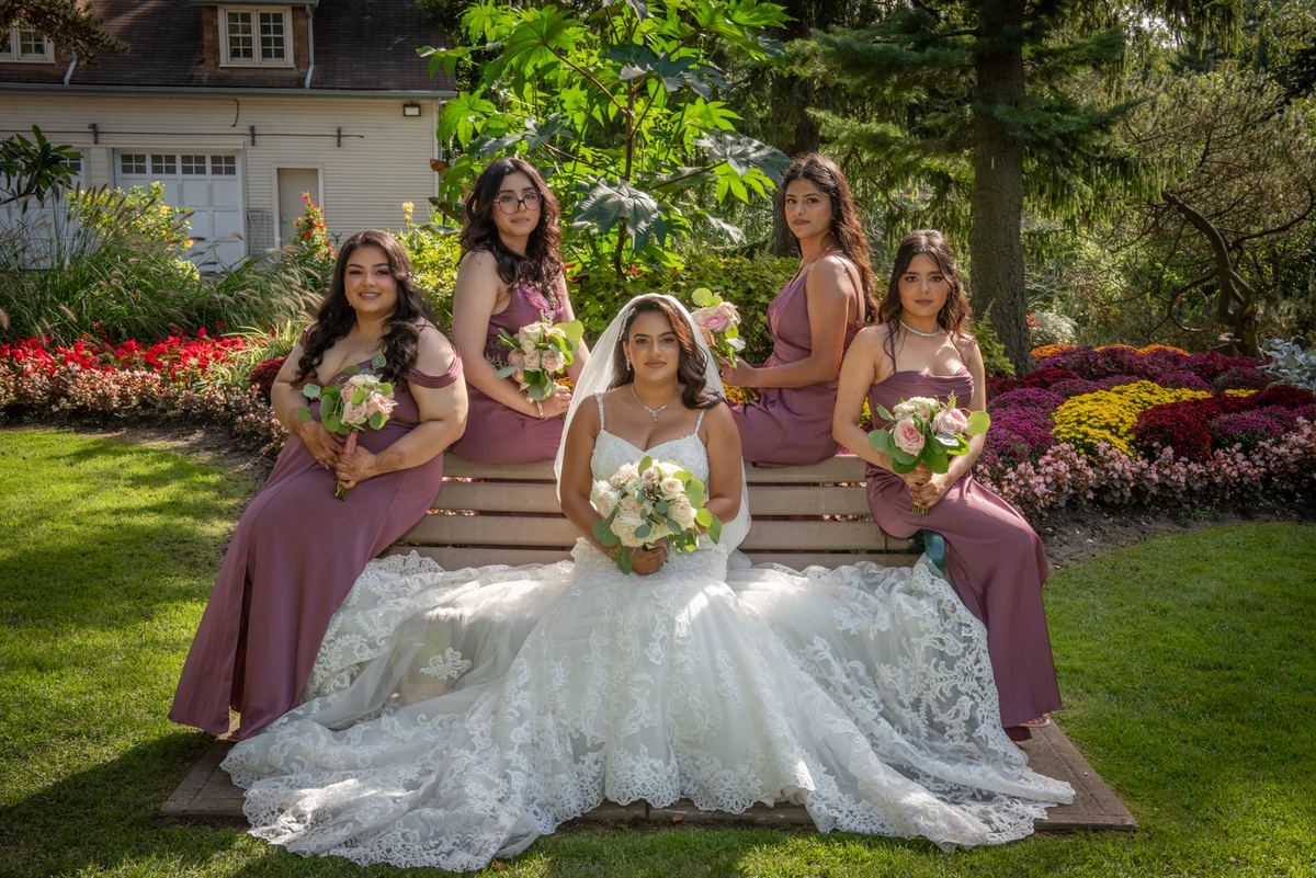 A bride in a white lace gown sits on a wooden bench with four bridesmaids in mauve dresses, surrounded by sunlight.