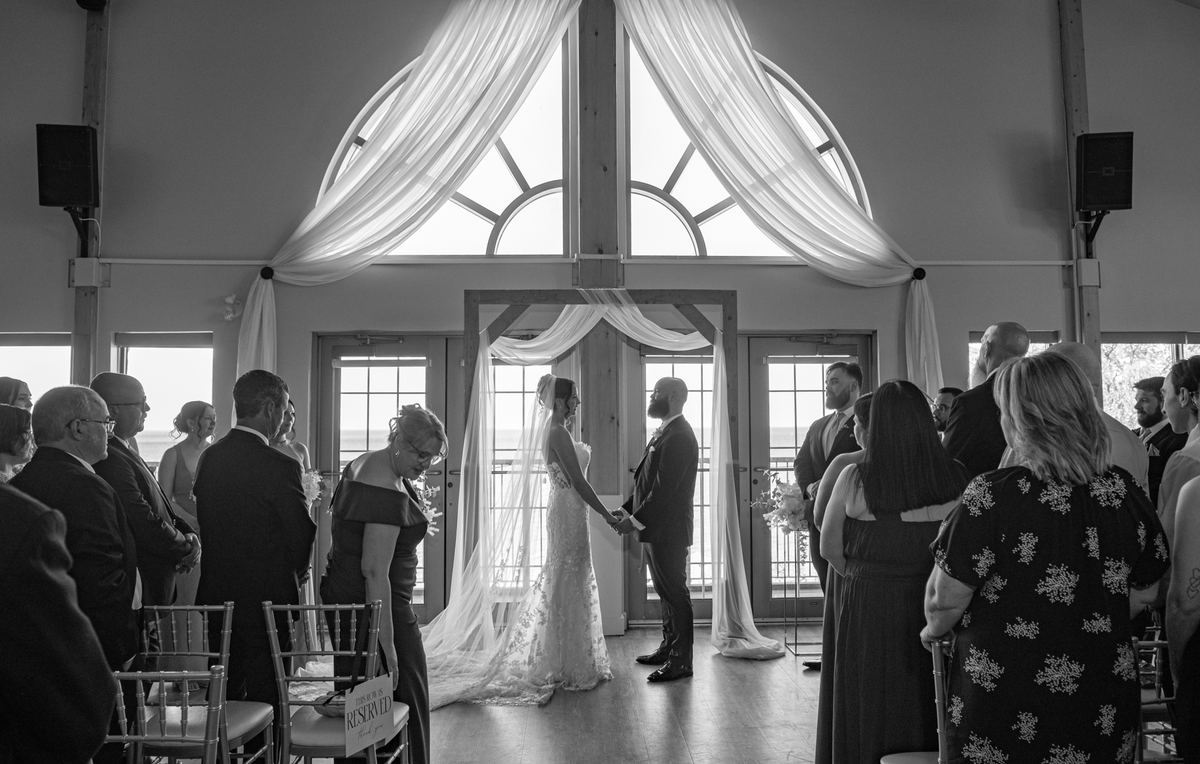 Bride and groom stand at an altar, hands clasped, framed by a sheer-draped window in soft light during the ceremony.