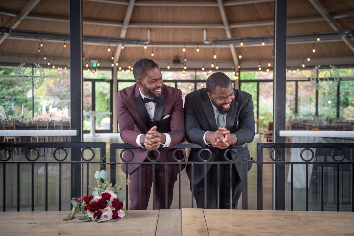 Two men in formal suits laugh by a railing, surrounded by fairy lights and a floral arrangement on a wooden table.