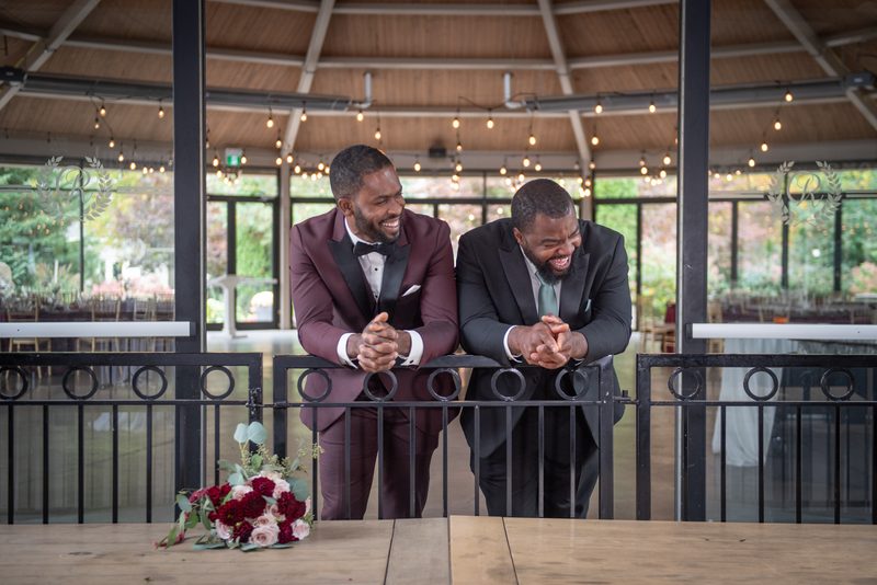 Two men in formal suits laugh by a railing, surrounded by fairy lights and a floral arrangement on a wooden table.