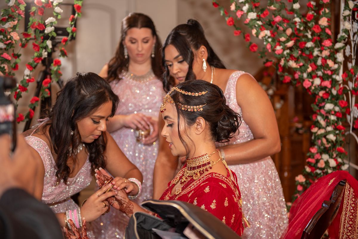 A bride in red and gold is seated as attendants apply henna to her hands in an intimate setting with floral arches.