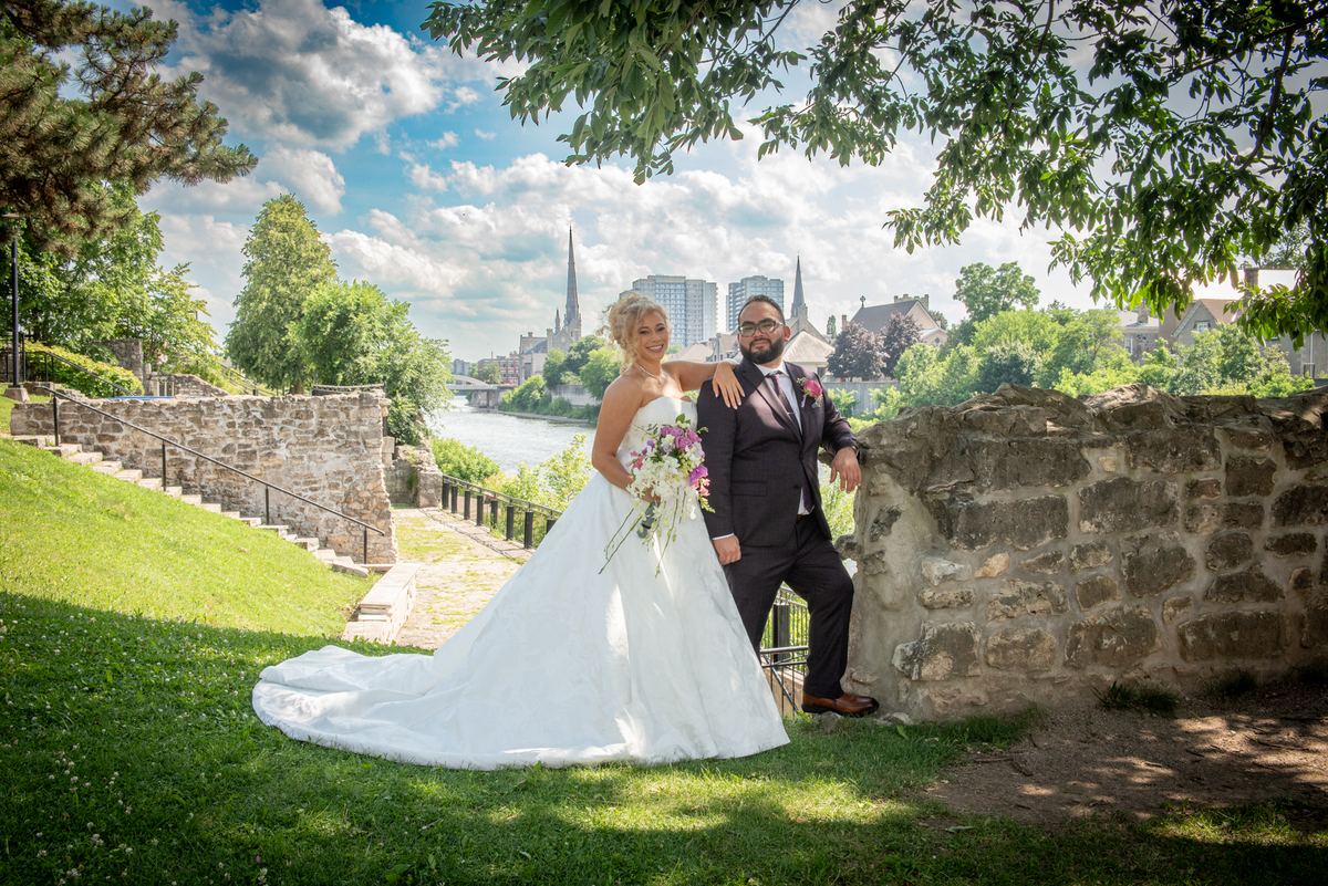 A bride in a white gown and groom in a dark suit pose joyfully beside a stone wall with a scenic cityscape behind them.