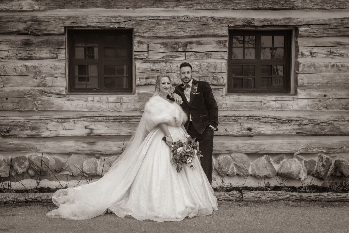 A bride in a voluminous gown and fur stole stands with her groom beside a rustic log cabin, the scene captured in monochrome.