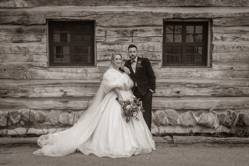 A bride in a voluminous gown and fur stole stands with her groom beside a rustic log cabin, the scene captured in monochrome.