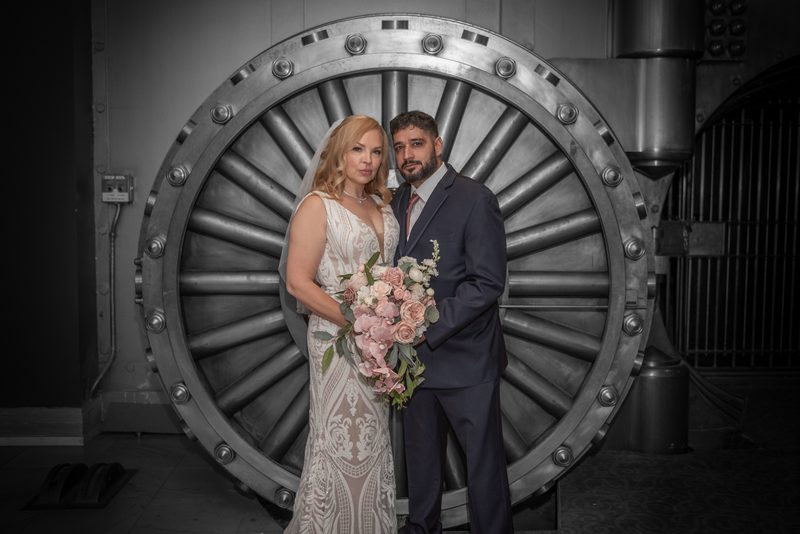 A bride and groom stand before an industrial metal vault door, contrasting the soft pink bouquet with the stark background.