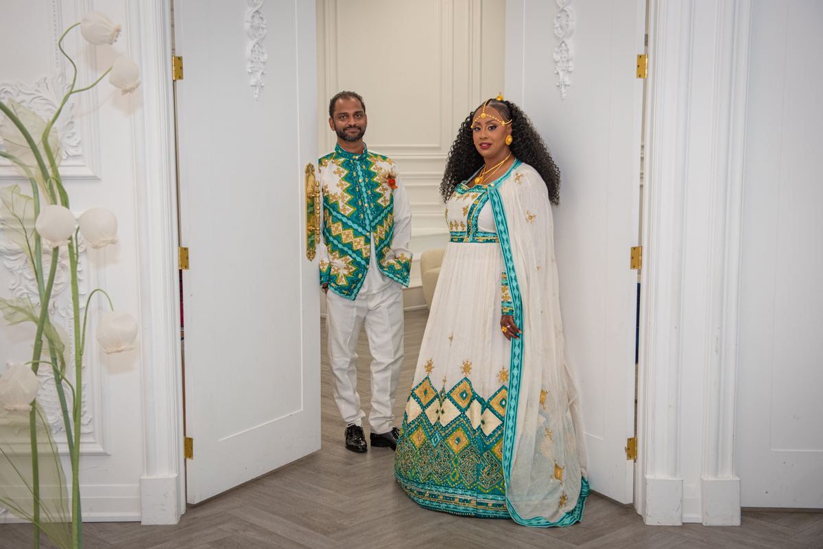 A couple in rich teal and gold ceremonial attire stands in an elegant doorway, illuminated by soft light.