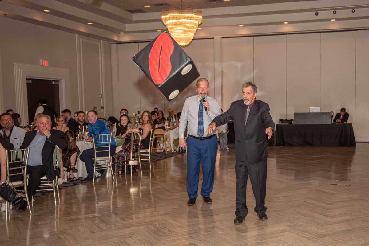 Two men perform with a suspended die at a banquet hall event, engaging guests with gestures and a microphone.