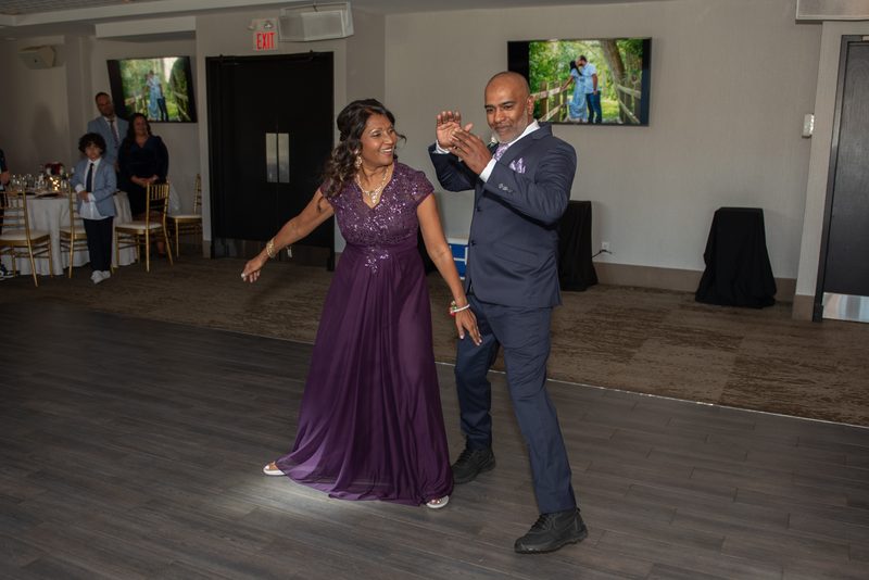 A smiling couple dances joyfully on a wooden floor under spotlights, surrounded by guests and framed photos.