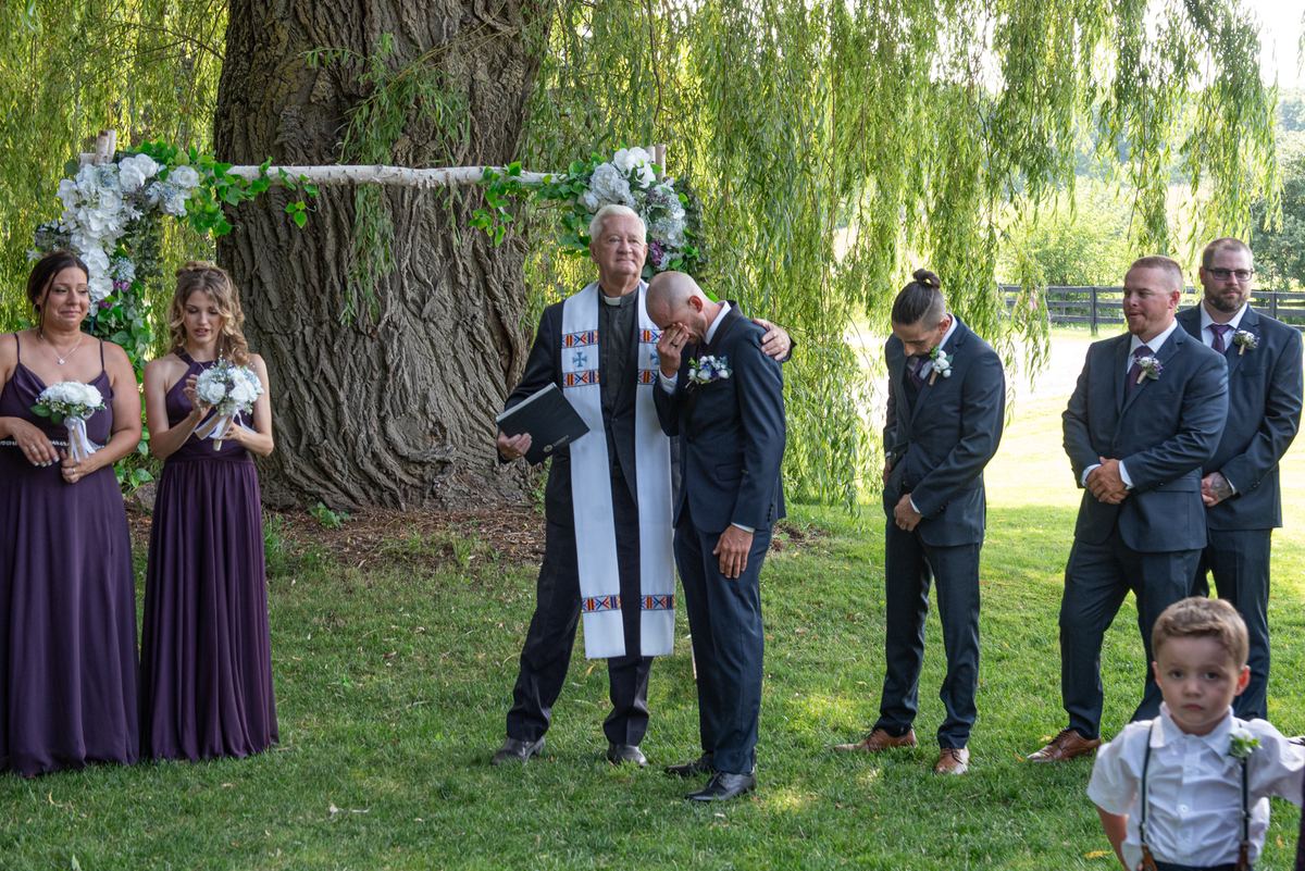 A groom covers his face as the officiant comforts him during an outdoor wedding under a willow tree.