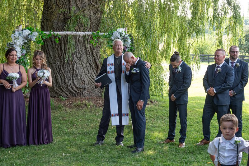 A groom covers his face as the officiant comforts him during an outdoor wedding under a willow tree.