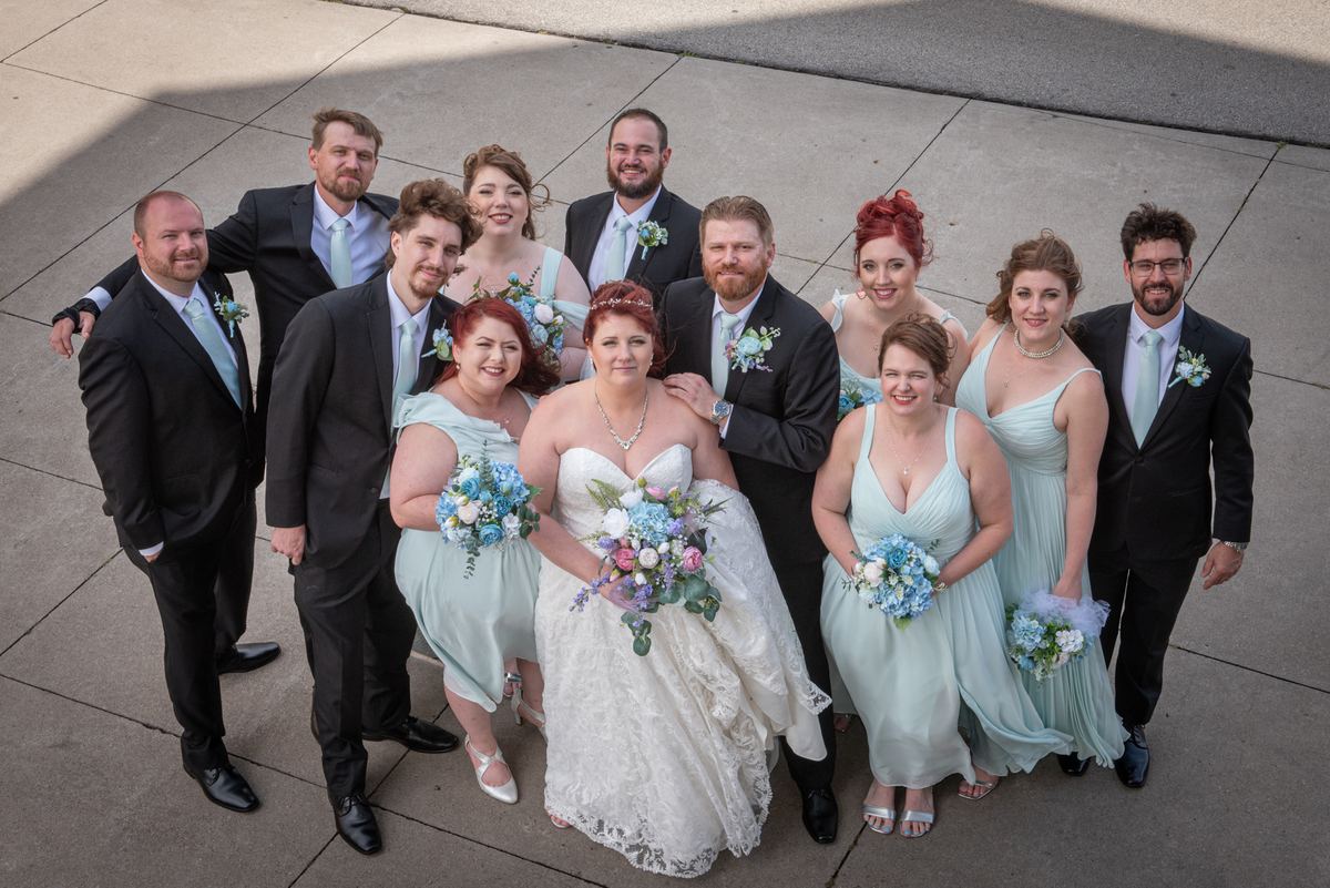Bride and groom stand with smiling bridesmaids in mint-green dresses and groomsmen in suits, under bright daylight.