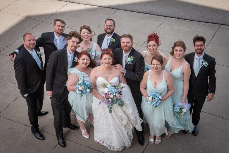 Bride and groom stand with smiling bridesmaids in mint-green dresses and groomsmen in suits, under bright daylight.