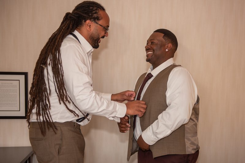 A man with dreadlocks adjusts the vest of his smiling companion, showing affection and anticipation.