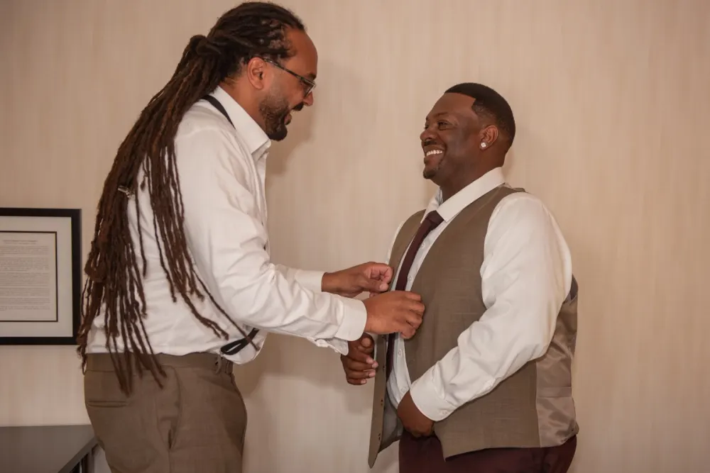 A man with dreadlocks adjusts the vest of his smiling companion, showing affection and anticipation.