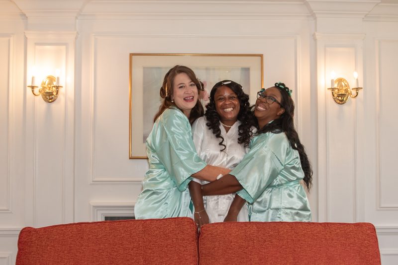 Three women in matching green and white robes laugh together in a warmly lit room with elegant white walls and red sofa.