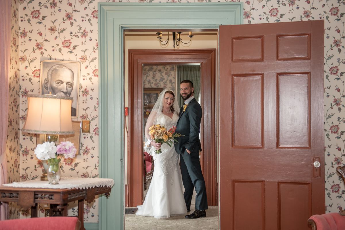 A bride and groom smile in a vintage doorway with floral wallpaper and an open wooden door, illuminated by soft light.
