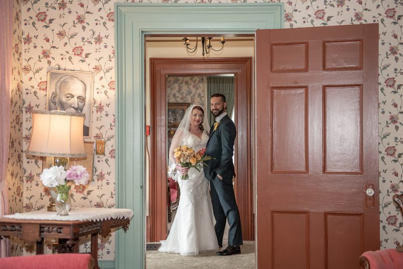 A bride and groom smile in a vintage doorway with floral wallpaper and an open wooden door, illuminated by soft light.