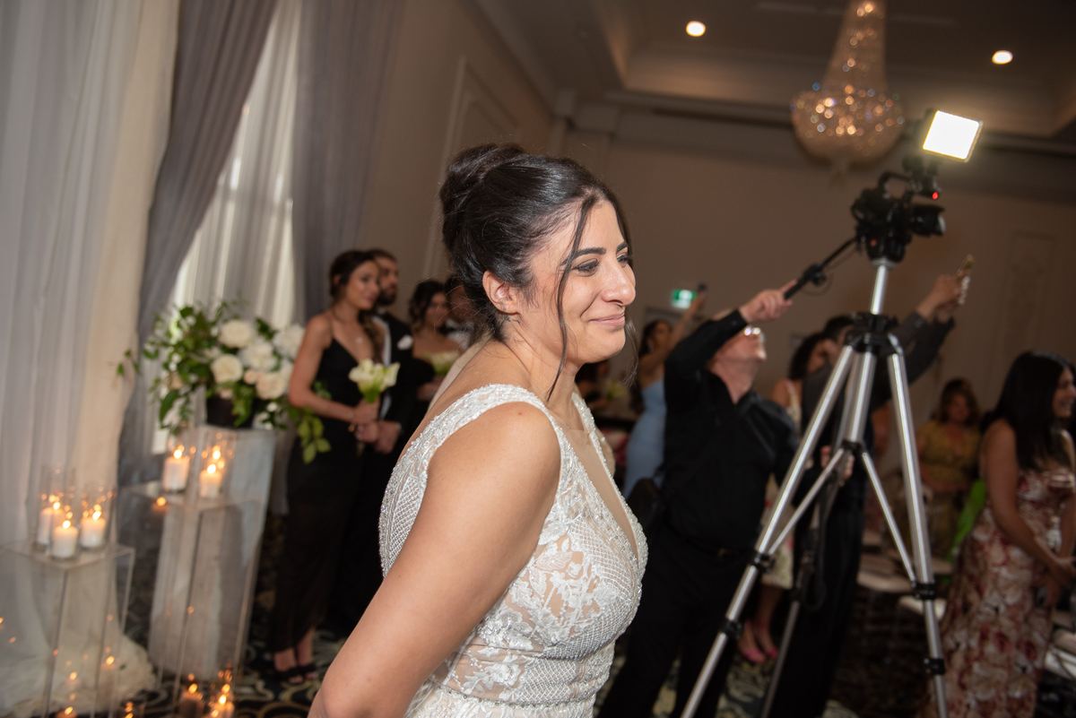 A smiling bride stands in a warmly lit reception room surrounded by guests and camera equipment, illuminated by candlelight.