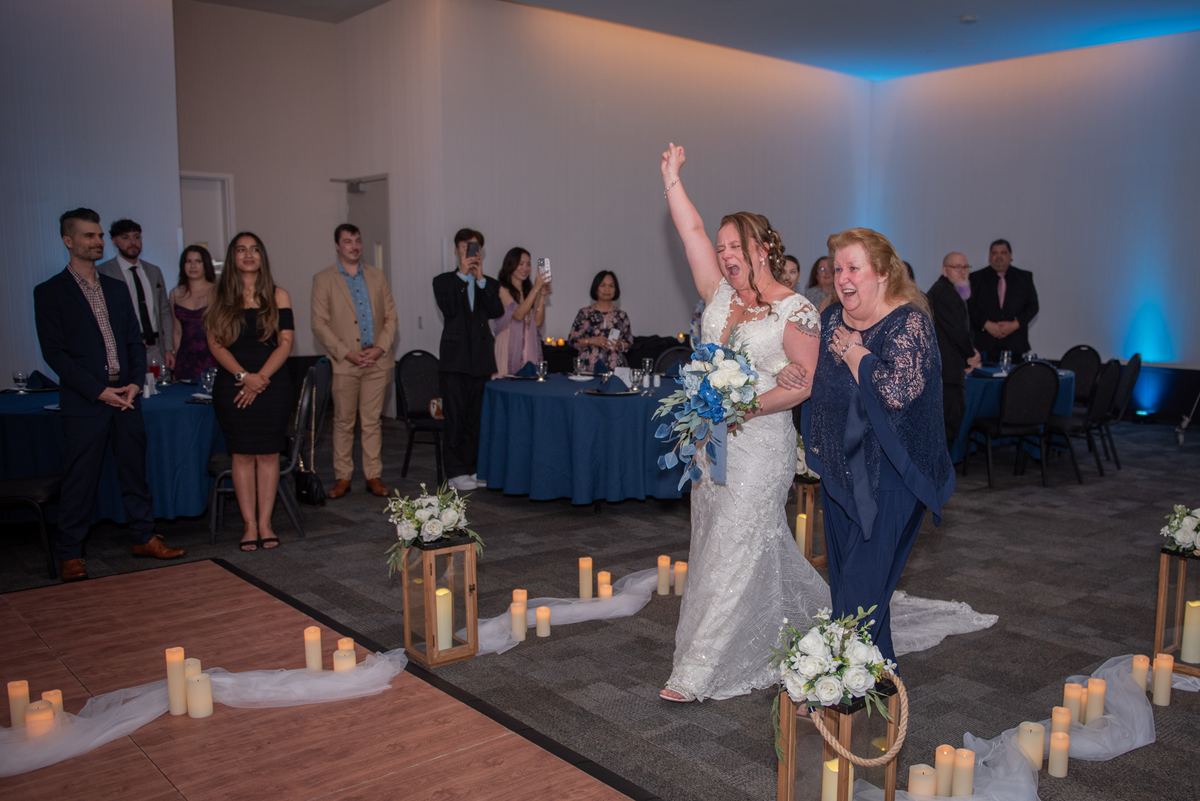 A bride in a white gown raises her arm triumphantly while walking down an aisle lined with candles and lanterns.