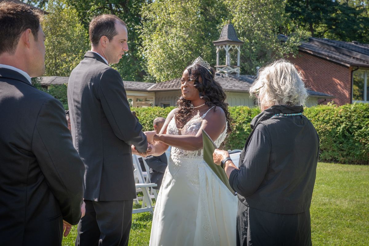 A bride and groom exchange vows under sunlight, hands clasped, with greenery and a brick building in the background.