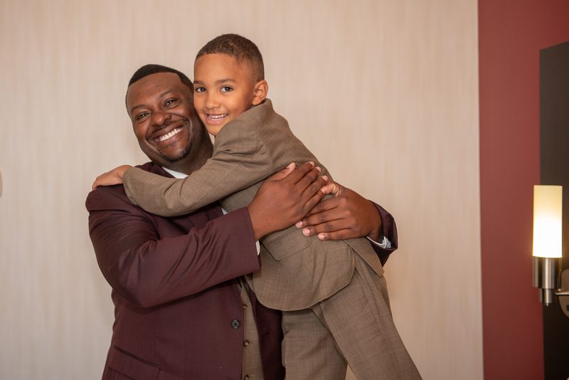 A man and a young boy in suits embrace warmly, smiling at the camera, illuminated by a modern wall sconce.