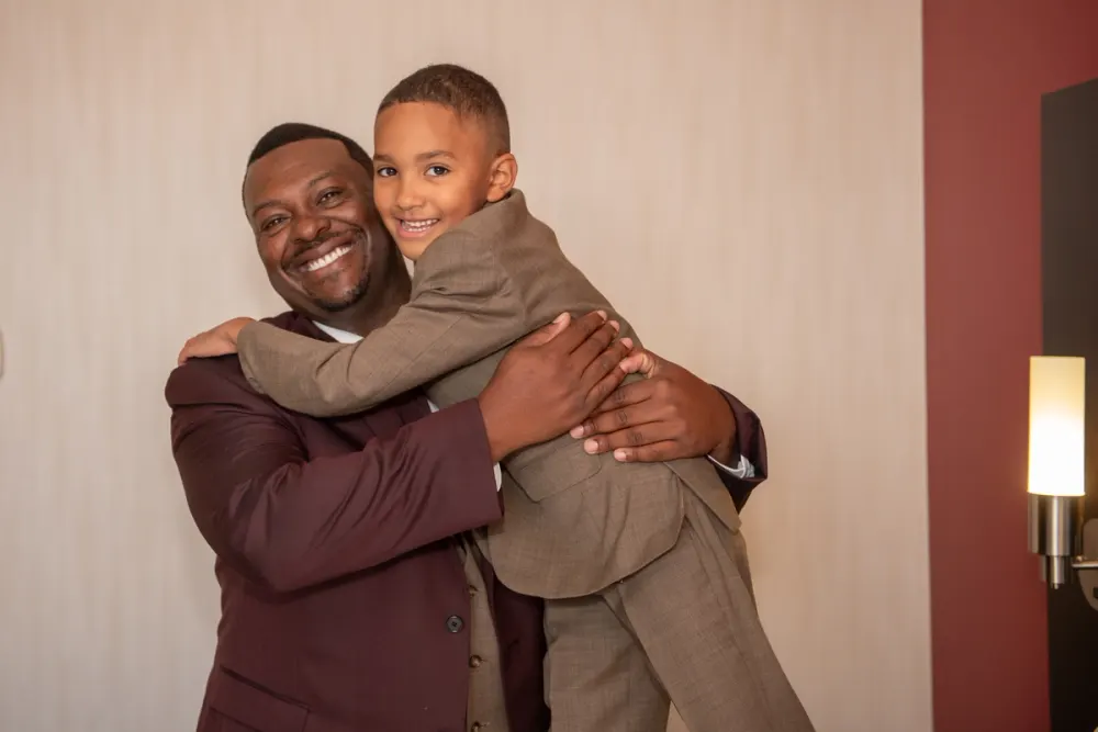 A man and a young boy in suits embrace warmly, smiling at the camera, illuminated by a modern wall sconce.