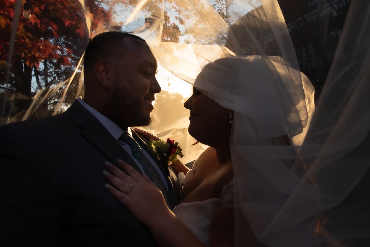 Bride and groom smile intimately, hands touching, as golden sunset light filters through a sheer veil.