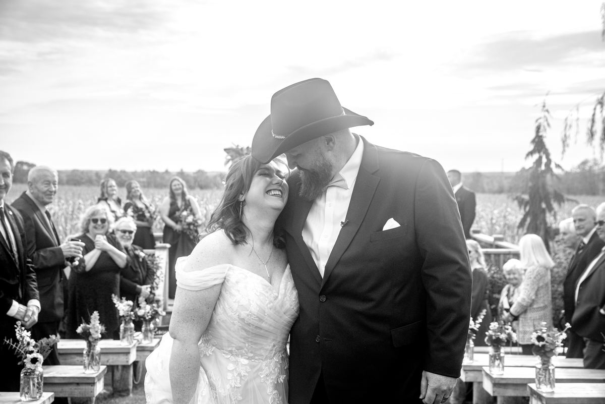 A laughing bride and groom share a kiss during an outdoor ceremony with guests watching from wooden benches.