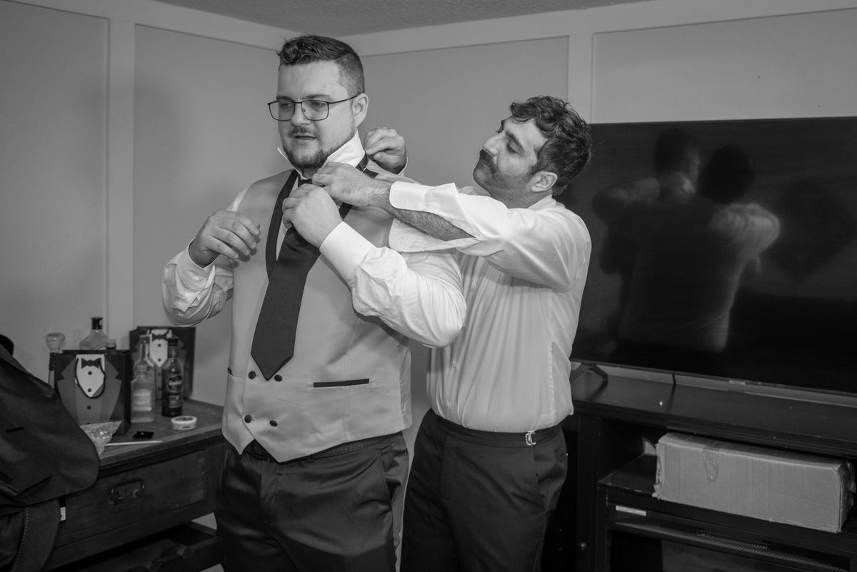 A man adjusts the tie of another in a softly lit room, with natural light highlighting their focused expressions.