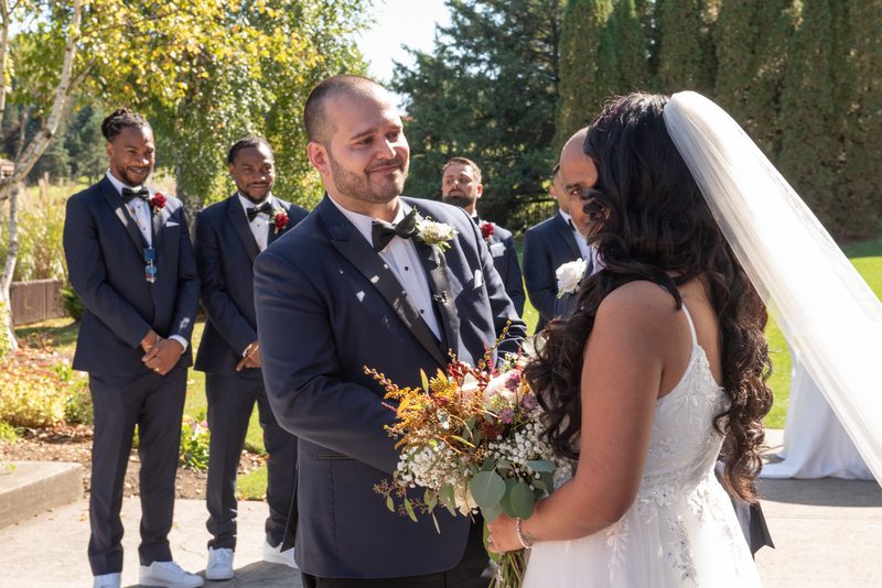 The groom smiles at his bride holding a colorful bouquet in an outdoor garden setting with trees and greenery.