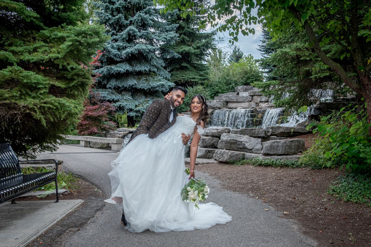 Bride and groom smile as he lifts her with a bouquet in hand near a stone waterfall surrounded by greenery.