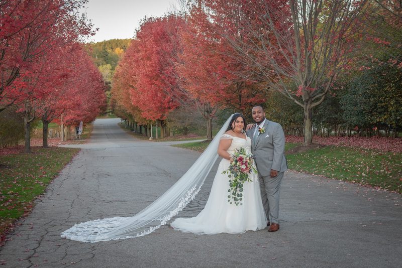 A bride in a long veil holding red flowers stands with her groom in a grey suit on a road lined with crimson autumn trees.