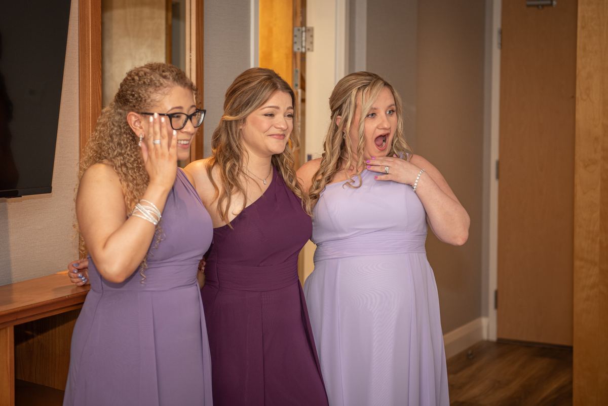 Three bridesmaids in pastel gowns express joy and surprise, their faces illuminated by soft indoor lighting.