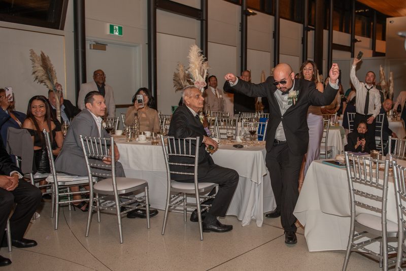 A groom in a black suit and sunglasses dances joyfully at a reception with guests watching and smiling.