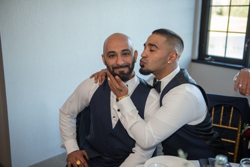 Two formally dressed men share an affectionate kiss, smiling joyfully as natural light highlights their elegant attire.