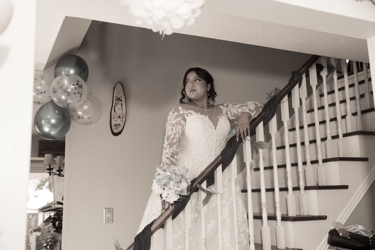 A bride in a lace wedding gown stands thoughtfully on a staircase, hand resting on the railing near festive balloons.