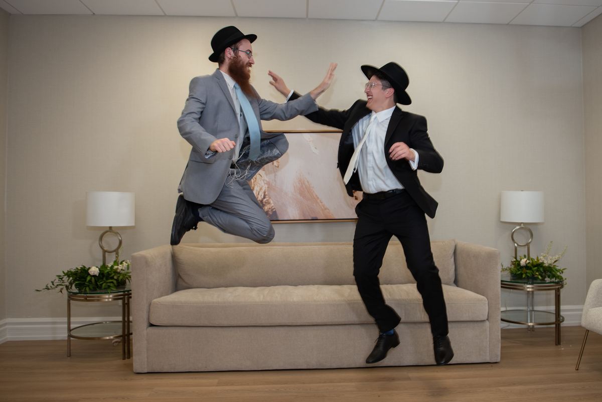 Two men in traditional Jewish attire leap joyfully into an embrace above a beige sofa in a brightly lit room.