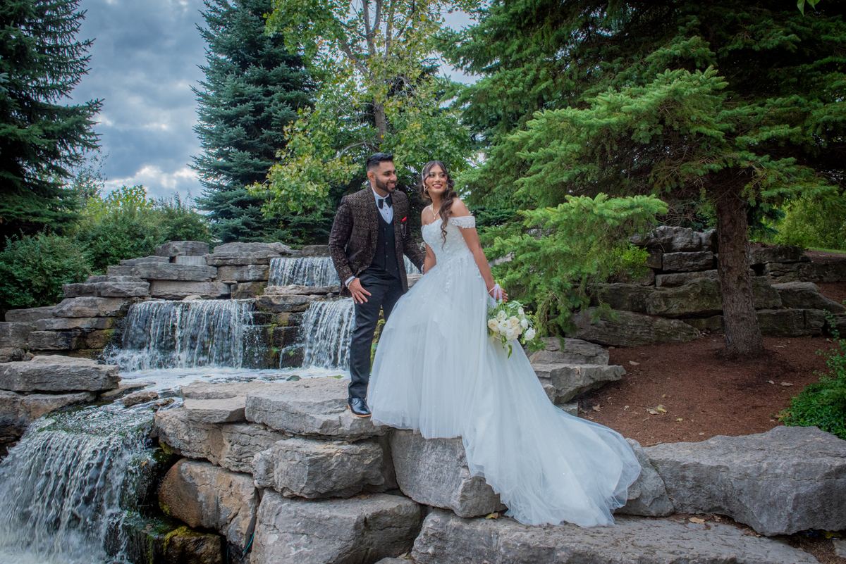 A bride in a white gown and groom beside a stone waterfall, framed by evergreen trees under a cloudy sky.